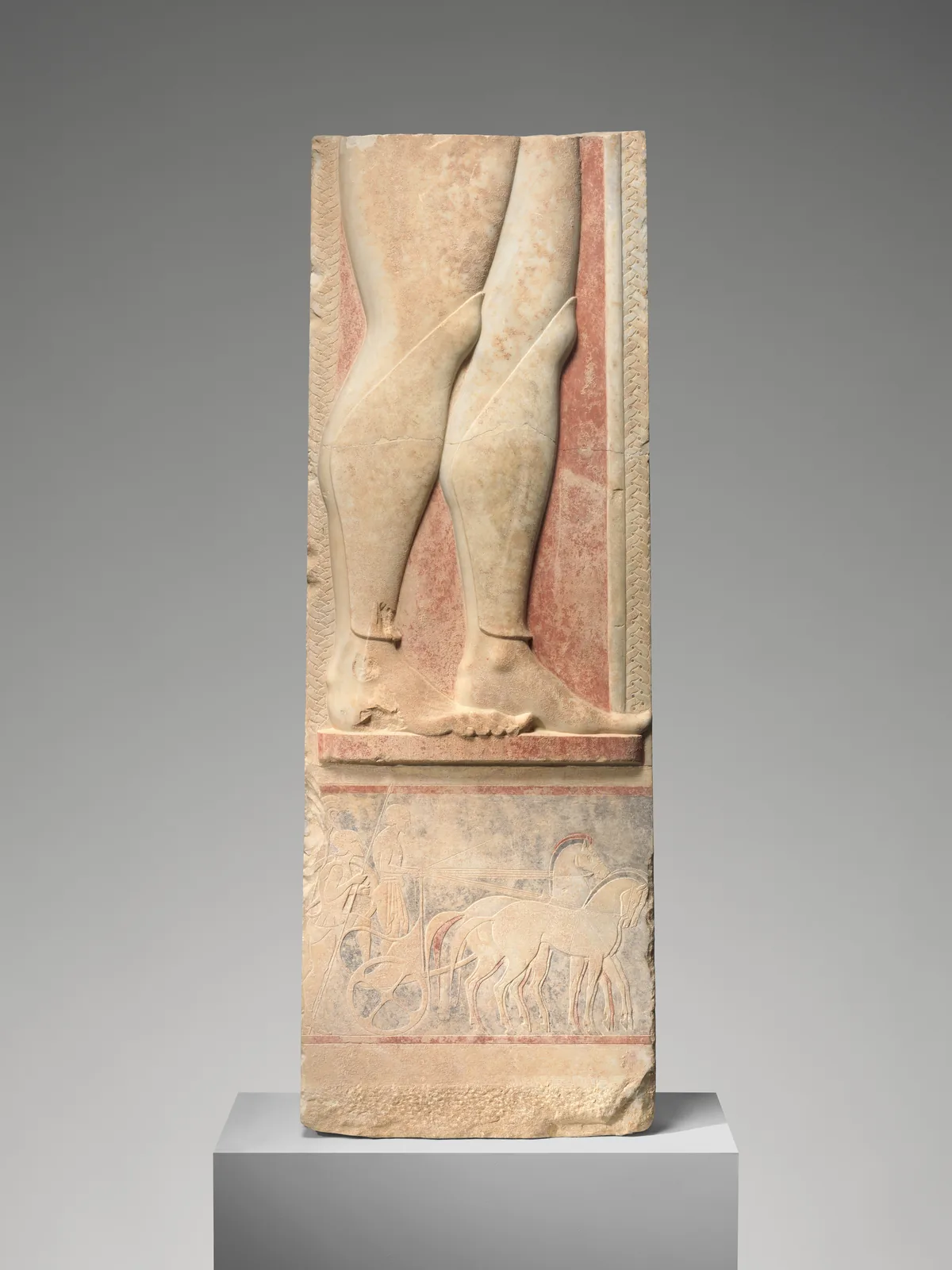 Fragment of an Attic marble grave marker showing a hoplite’s lower legs and a chariot scene.