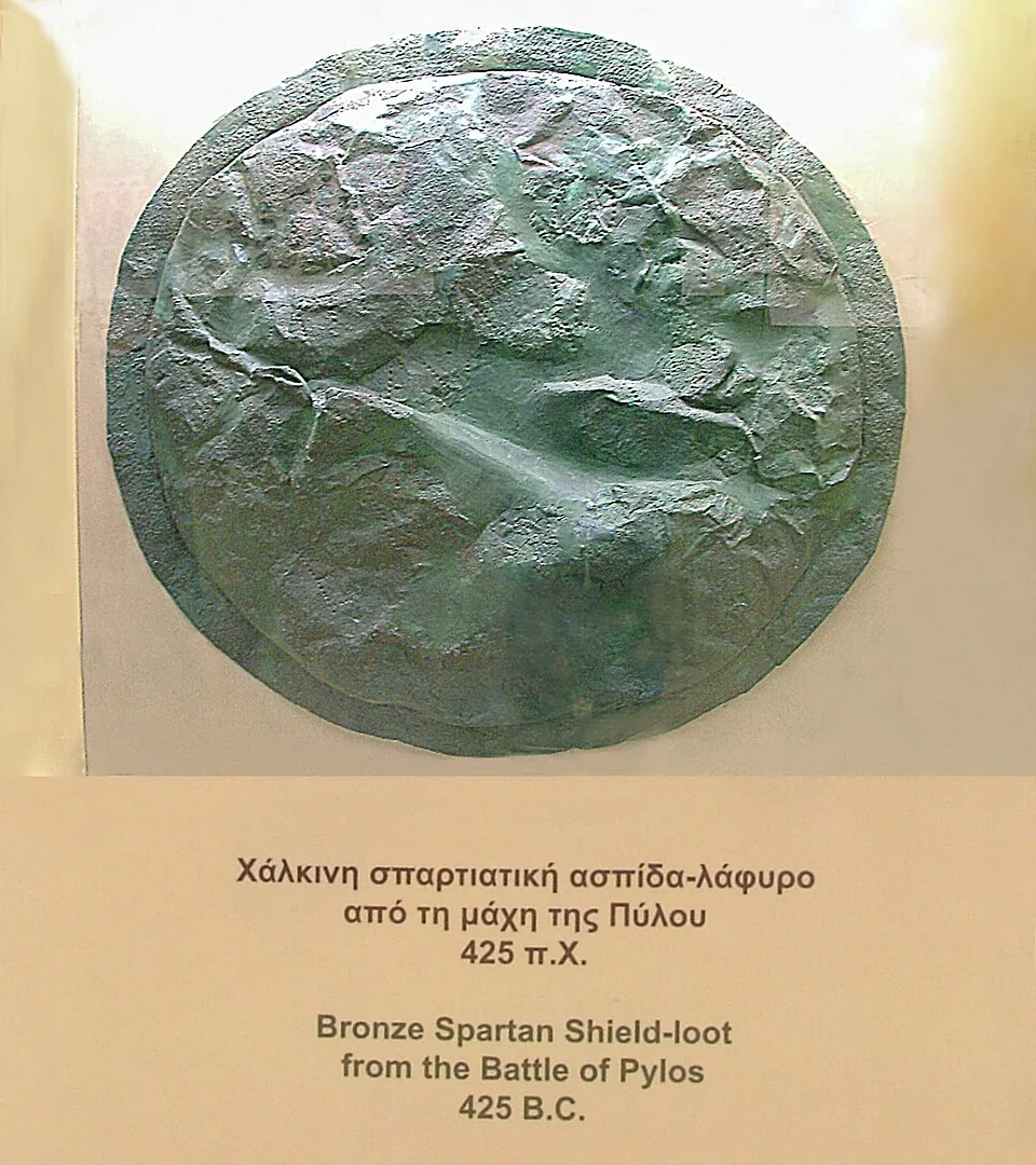 A battered bronze Spartan shield captured at Pylos, bearing a punched inscription.